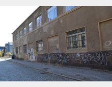 Straßenszene mit einem alten, beige-braunen Gebäude aus verputztem Mauerwerk. Die Fassade zeigt Spuren von Verfall, Graffiti und vergitterte Fenster im Erdgeschoss; der Gehweg und die Straße sind gepflastert.