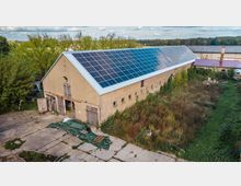 Ein längliches, einstöckiges Gebäude mit hellem Putz und mehrfachen kleinen Fenstern, dessen Dach fast vollständig mit Solarpaneelen bedeckt ist. Das Gebäude steht in einer teils betonierten, teils überwucherten Umgebung mit Büschen und Gras im Hintergrund.