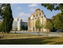 Ruine eines historischen Backsteingebäudes mit großen, runden Fensteröffnungen, umgeben von Bäumen und einer Grünfläche, daneben ein modernes mehrstöckiges Bürogebäude in heller Fassade.