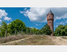 Ein historischer roter Backsteinturm mit spitzem Schieferdach steht auf einem Hügel, umgeben von Bäumen und Gebüsch. Im Vordergrund sind ein sandiger Weg und ein Zaun mit Holzpfählen zu sehen, dahinter blauer Himmel mit weißen Wolken.