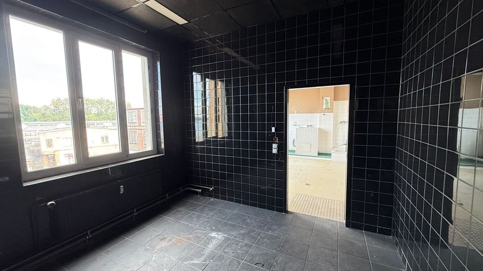 Leerer Innenraum mit schwarz gefliesten Wänden und Boden, abgehängter Decke mit Leuchtstoffröhre und großem Fenster zur Außenansicht. Rechts führt eine Türöffnung in einen helleren, ebenfalls gefliesten Nebenraum.