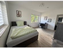 Modern eingerichtetes Schlafzimmer mit hellem Holzfußboden. Ein graues Polsterbett mit grüner Bettwäsche steht links neben einem Fenster, rechts befindet sich ein Schreibtisch mit Stuhl.