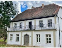 Weiß gestrichenes zweistöckiges Gebäude mit rotem Ziegeldach, symmetrischen Fenstern und einem zentralen, grauen Eingang mit halbrundem Bogen. Auf der oberen Etage befindet sich ein schmiedeeisernes Geländer, daneben ein großer Baum und eine natürliche Umgebung im Hintergrund.