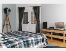 Schlafzimmer mit Holzboden und einer karierten Decke auf dem Bett. Im Raum befinden sich eine Stehlampe im Vintage-Stil, ein Holzregal mit Schallplattenspieler und Lautsprecher sowie eine Tür mit Glaseinsätzen, die von grünen Vorhängen umrahmt wird.