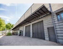 Einfache Industrie- oder Lagergebäude-Fassade mit grauen Metalltoren und -türen, einem Vordach aus Metall und einer gepflasterten Vorfläche; daneben Bäume, geparkte Fahrräder und Autos.