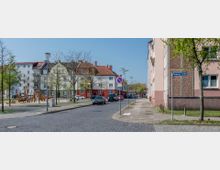 Straßenszene in einem Wohngebiet mit mehrstöckigen Häusern im klassischen Stil, teilweise mit roten Dächern. Auf der linken Seite befindet sich ein kleiner Spielplatz mit Holzspielgeräten, während Bäume und parkende Autos die Straße säumen.