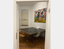 Ein kleiner, heller Raum mit Holzboden, einem schwarzen rechteckigen Tisch auf Metallbeinen und verschiedenen Stühlen. An der Wand hängt ein farbenfrohes Gemälde, und im Hintergrund ist eine hölzerne Tür mit vergittertem Oberlicht sichtbar.