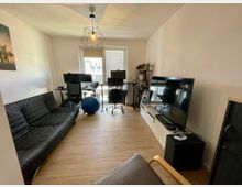 Wohnzimmer mit multifunktionalem Design: Im Vordergrund steht ein schwarzes Ledersofa, rechts ein großer Flachbildfernseher auf einem weißen TV-Schrank. Im hinteren Bereich befinden sich zwei Schreibtische mit Monitoren und Bürostühlen, daneben ein blauer Gymnastikball. Der Raum hat Holzboden und wird durch ein Fenster mit Tageslicht erhellt.