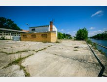 Industriegelände mit leerer Betonfläche, einem kleinen Backsteingebäude und einem größeren weißen Gebäude im Hintergrund; rechts verläuft ein Fluss mit Geländer entlang der Uferseite, umgeben von Bäumen und Vegetation.