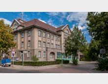 Eckgebäude im Stil der frühen Moderne mit vier Stockwerken, heller Fassade und rotem Satteldach, umgeben von Bäumen und Straßen in einer verkehrsberuhigten Zone. Vor dem Gebäude stehen Verkehrsschilder und ein blauer Anhänger.