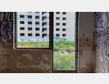 Blick aus einem verlassenen, mit Graffiti bedeckten Raum durch ein offenes Fenster auf ein leerstehendes Betongebäude mit vielen kleinen Fensteröffnungen. Im Vordergrund ist eine verwilderte Fläche mit Gras und gelbem Unkraut zu sehen, die teilweise mit Graffiti versehen ist.