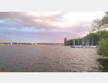 Ein See mit ruhiger Wasseroberfläche, umgeben von Bäumen. Rechts am Ufer liegen zahlreiche Segelboote an einem kleinen Steg, im Hintergrund sind Gebäude und eine bewaldete Küstenlinie sichtbar.