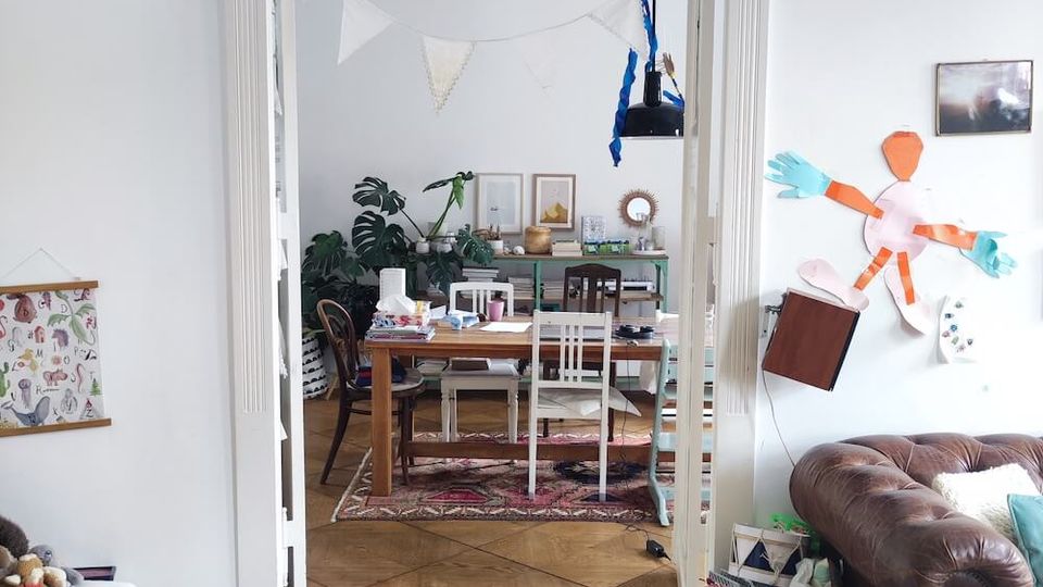 A cozy living and dining space with light-colored walls, wooden floors, and an arched door frame separating the rooms. The dining area features a wooden table and chairs with potted plants and framed art in the background, while the living space includes a leather sofa and scattered toys and decorations.