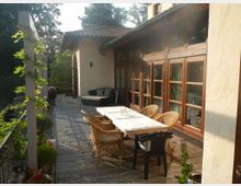 Eine Terrasse mit Holzboden, an der ein rechteckiger Tisch mit Stühlen aus Rattan und Kunststoff steht. Im Hintergrund steht ein Gebäude mit großen Holztüren und Fenstern, flankiert von Pflanzen und Pergolaelementen; umliegende Bäume grenzen die Terrasse ein.