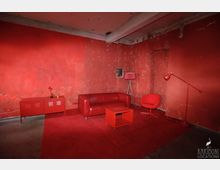 Ein industrieller Raum mit abgenutzten roten Wänden, einer roten Couch, einem roten Teppich und roten Möbeln wie einem Beistelltisch, einem Sideboard sowie einer Steh- und Tischlampe. Die Backsteinstruktur der Wände ist sichtbar, und die Decke zeigt rohe Betonbalken.