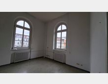 Leerer, heller Innenraum mit weißen Wänden und zwei großen, rundbogigen Sprossenfenstern, darunter jeweils ein Heizkörper. Grauer Boden und sichtbare Leitungen entlang der Wand; durch die Fenster sind gegenüberliegende Wohngebäude zu sehen.