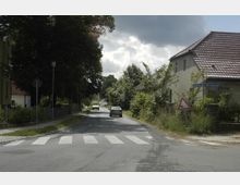 Eine ländliche Straße mit Zebrastreifen, flankiert von alten Häusern mit roten Ziegeldächern und dichtem Bewuchs. Straßenschilder zeigen die Richtung zu „Neu-Langerwisch“, und ein Verkehrsschild warnt vor einer Steigung; im Hintergrund fahren ein gelber und ein dunkler PKW.