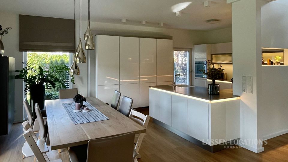 Heller, moderner Küchen- und Essbereich mit großem Holzesstisch, Stühlen und hängenden Glas-Pendelleuchten. Dahinter eine weiße, grifflos gestaltete Einbauküche mit Kücheninsel und dunkler Arbeitsplatte, neben großen Fenstern mit Blick ins Grüne.