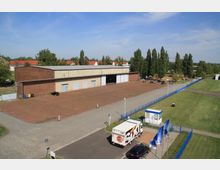 Industriehalle aus Backstein und Beton mit großem gepflasterten Vorplatz, daneben eine Rasenfläche, die durch einen blauen Zaun abgegrenzt ist. Im Vordergrund parkt ein Lieferwagen vor einem kleinen Wachhäuschen, und vereinzelte Autos stehen am Gelände verteilt.