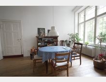Wohnzimmer mit Holzboden, einem runden Tisch mit blau-weiß gepunkteter Tischdecke und fünf Holzstühlen. Im Hintergrund stehen ein Bücherregal, Pflanzen in Töpfen sowie ein großes Fenster, das Licht in den Raum lässt.