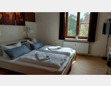 Ein schlicht eingerichtetes Schlafzimmer mit einem Doppelbett, frischer Bettwäsche und gefalteten Handtüchern. Im Hintergrund befindet sich ein großes Fenster mit braunen Vorhängen, das Tageslicht hereinlässt, sowie eine helle Wand mit einer Weltkarten-Dekoration.