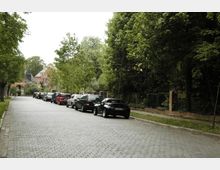 Gepflasterte Straße in einer grünen Wohngegend, gesäumt von geparkten Autos auf der linken Seite und einem Zaun mit Bäumen auf der rechten Seite. Im Hintergrund sind rote Backsteinhäuser mit Spitzdach und umliegende Vegetation sichtbar.