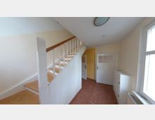 Treppenhaus mit einer weißen Holztreppe und einem Holzgeländer, dahinter zwei weiße Türen mit Glasausschnitten und Wand in beige und gelber Farbe. Der Boden ist mit braunen Fliesen ausgelegt, ein Fenster und ein Heizkörper befinden sich auf der rechten Seite.