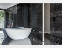 Modernes Badezimmer mit freistehender, weißer Badewanne auf glänzendem dunkelgrauem Marmorboden. Die Wände sind ebenfalls dunkelgrau verkleidet, und durch ein großes Fenster fällt natürliches Licht herein.