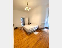 Schlafzimmer mit Holzboden und weiß gestrichenen Wänden. Ein Doppelbett in der Mitte des Raumes steht auf einem hellen Holzfußboden; daneben ein Nachttisch mit Lampe und Bücherstapel. Im Hintergrund befindet sich eine geöffnete zweiflügelige Tür.