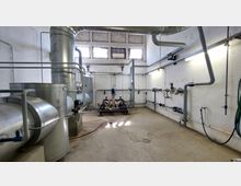 Industrieanlage mit großen Lüftungsrohren, Ventilen und Rohrleitungen in einem weißen Raum mit hohen Decken. Tageslicht fällt durch mehrere kleine Fenster an der Oberseite der Wand ein, ergänzt durch künstliche Beleuchtung.