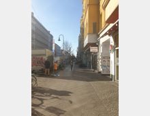 Eine städtische Straße mit Kopfsteinpflaster, flankiert von Gebäuden mit Läden und Restaurants, darunter ein Schild mit der Aufschrift "Steinofen Pizza". Im Hintergrund sind Fahrräder, ein Lieferwagen und Passanten zu sehen, bei klarem Himmel und Sonnenlicht.