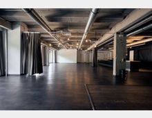 Großer, offener Innenraum mit industriellem Design, sichtbaren Lüftungsrohren an der Decke, grauem Bodenbelag und schwarzen Vorhängen an den Seiten. Der Raum enthält massive Betonstützen, eine Bar auf der rechten Seite und große Fenster, die für Tageslicht sorgen.