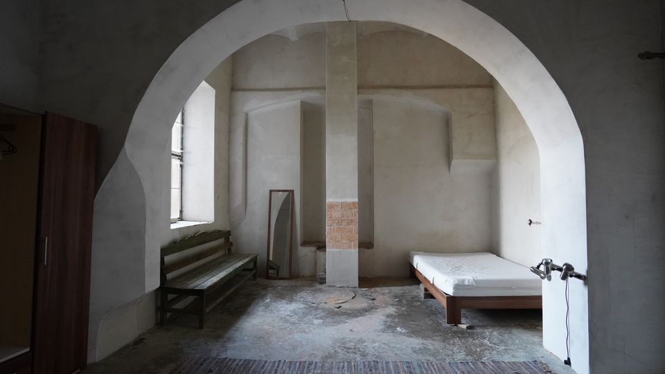 Spärlich eingerichteter, karger Innenraum mit großem Rundbogen, verputzten Wänden und rohem Betonboden, in der Mitte von einer massiven Stütze geteilt. Links stehen eine Holzbank und ein Fenster, rechts ein einfaches Bett; außerdem sind ein Spiegel und ein Schrank zu sehen.