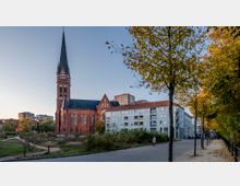 Rote Backsteinkirche mit hohem, spitzen Turm und großen gotischen Fenstern, umgeben von Grünflächen und Bäumen in herbstlichen Farben. Im Hintergrund sind moderne Wohngebäude zu sehen, rechts verläuft eine Gehwegallee mit Baumreihe.