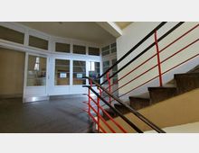 Treppenhaus in einem öffentlichen oder Wohngebäude mit einer Stein- oder Betontreppe, schwarzem Handlauf und roten Geländerstreben. Im Hintergrund sind weiße Türen mit großen Glaseinsätzen zu sehen, die einen weiteren Raum abschließen.