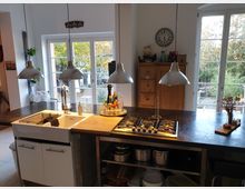 Offene Küche mit einer zentralen Kücheninsel, die eine Spüle, ein Kochfeld und Hängelampen umfasst. Im Hintergrund sind große Fenster und Glastüren zu einem Garten mit herbstlicher Vegetation sichtbar. Ein Holzschrank und Küchenutensilien ergänzen die Einrichtung.