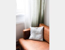 Braunes Ledersofa mit einem weißen Kissen, daneben ein Fenster mit halbtransparenten weißen und grünen Vorhängen; im Hintergrund befindet sich ein Heizkörper.