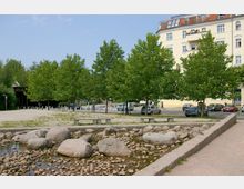 Öffentlicher Platz mit gepflastertem Bereich und einem kleinen, steinigen Wasserlauf im Vordergrund, flankiert von Bänken. Im Hintergrund befinden sich dicht bepflanzte Bäume, parkende Autos und ein mehrstöckiges Wohngebäude mit gelber Fassade.
