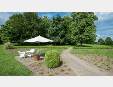Grüne Parklandschaft mit einem gepflasterten Weg, der durch gepflegte Beete und Rasenflächen führt. Im Vordergrund steht eine Sitzgruppe mit weißen Stühlen und einem Tisch unter einem Sonnenschirm, umgeben von hohen Bäumen.