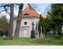 Kleine Kapelle mit rosa und weißen Wänden sowie einem roten Ziegeldach, umgeben von Bäumen, Grabsteinen und Rasen. Im Vordergrund stehen mehrere Kreuze auf einem Friedhof.