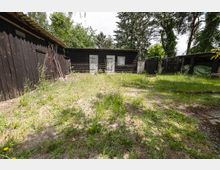 Innenhof mit unebenem, teils sandigem Boden und spärlichem Grasbewuchs, umgeben von dunklen Holzschuppen mit Wellblechdächern. Im Hintergrund stehen zwei helle Holztüren und ein kleines Fenster, daneben Zäune und hohe Bäume.
