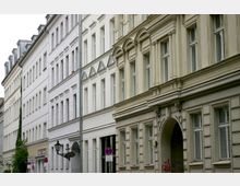 Straßenansicht mit historischen Gebäuden aus der Gründerzeit, gekennzeichnet durch hohe Fenster, Fassadendekorationen und einen Rundbogen-Eingang. Die Häuserwände sind größtenteils in hellen Farbtönen gehalten.