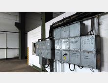 Metallene Schalttafeln und Verkabelungen an einer weißen Wand in einer industriellen Halle. Der Raum hat eine klare, funktionale Einrichtung mit Betonboden und sichtbaren Trägern.