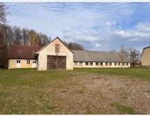 Ländlicher Bauernhof mit hellem Hauptgebäude und großer Scheunentür sowie einem langen, niedrigem Stall- oder Wirtschaftsgebäude mit vielen Fenstern. Davor liegt eine weite Wiese, im Hintergrund stehen kahle Bäume unter einem teils bewölkten Himmel.