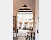 Heller Innenraum eines Cafés mit einer spitzbogigen Öffnung, die in einen weiteren Bereich mit langen Holztischen und Stühlen führt. An den Wänden hängen moderne Bilder, und von der Decke hängen Leuchten sowie Pflanzen in Töpfen.