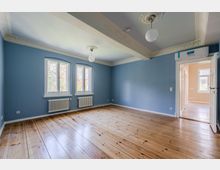 Leerer Wohnraum mit blau gestrichenen Wänden, breiten Holzdielen und zwei hohen Fenstern über weißen Heizkörpern. Stuckverzierte Decke mit Kugelleuchten und eine Türöffnung führt in einen hellen Nebenraum.