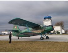 Ein grünes Doppeldecker-Flugzeug steht auf einer asphaltierten Startfläche vor einem kleinen Kontrollturm mit blauer Kuppel. Im Vordergrund ist eine Person auf einer Rasenfläche zu sehen, während im Hintergrund weitere Gebäude des Flughafens sichtbar sind.