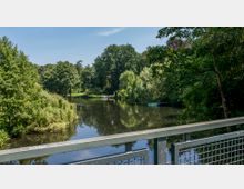 Blick von einer Brücke auf einen ruhigen Fluss umgeben von dichter Vegetation, darunter große Bäume und Weiden, mit vereinzelten Booten am Ufer. Im Hintergrund befindet sich eine grüne Parklandschaft unter einem klaren Himmel.