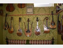 Ein Raum mit grün gemusterter Tapete, in dem eine Sammlung von antiken Saiteninstrumenten, darunter Mandolinen und Lyra-ähnliche Instrumente, an der Wand hängt. Darunter steht ein gepolstertes Möbelstück mit gestreiftem Stoffbezug.