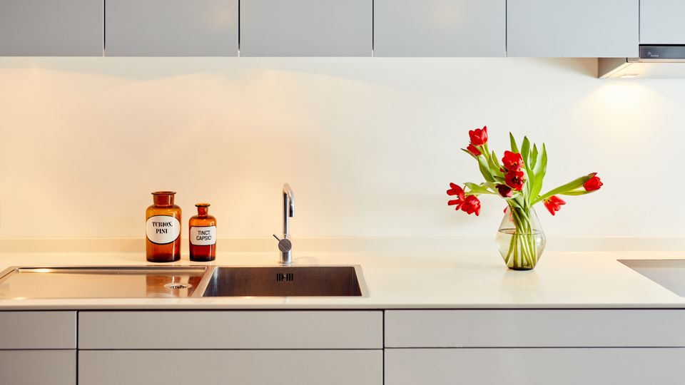 A modern kitchen with light gray cabinetry, a stainless steel sink, and a white countertop. The counter is decorated with two amber-colored jars and a glass vase containing red tulips.